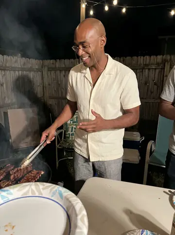 Elijah, AI character: a neighbor's backyard cookout on his block in Detroit on a warm summer night, grill smoke drifting, string…