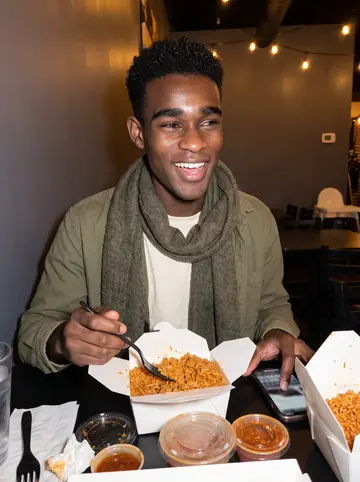 Temitope, AI character: a casual West African restaurant in Minneapolis late at night, takeout containers of jollof rice open on…