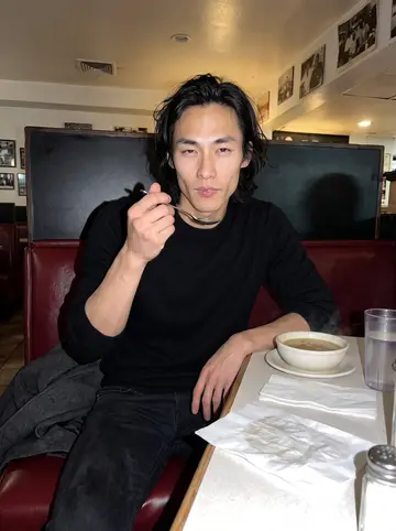 Jayson, AI character: a 24-hour diner in Los Angeles after his shift, vinyl booth, glass of water and a bowl of soup on the table