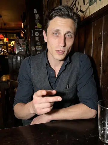 Maciej, AI character: a small low-ceiling bar near Sternschanze after work, dark wood wall and half-empty glasses behind him