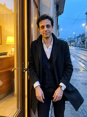 Anthony, AI character: outside the small hotel entrance near Grenoble station just after the night shift, blue streets behind him…