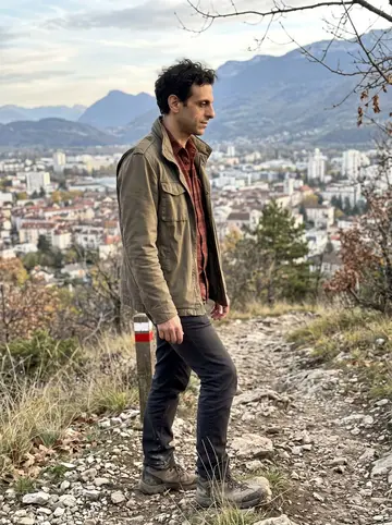 Anthony, AI character: a rocky path above Grenoble in the Chartreuse foothills, city roofs far below in the late autumn light
