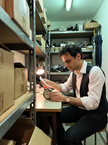 Anthony, AI character: the hotel service room after midnight, shelves of unclaimed chargers and umbrellas around a narrow work table