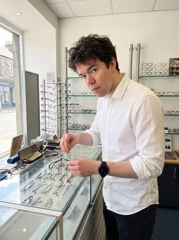 Sjoerd, AI character: inside the Inverness optician shop, beside a glass display counter and rows of eyeglass frames