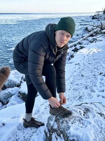 Nick, AI character: icy shoreline rocks near Lake Superior in January, snow crust along the path and blue water behind him